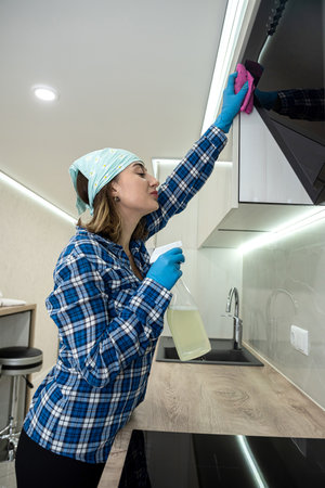 woman in a headscarf and gloves in the kitchen washes the kitchen set and the hood with a pink cloth. the concept of cleanliness in the kitchen. cleaning up after cookingの写真素材