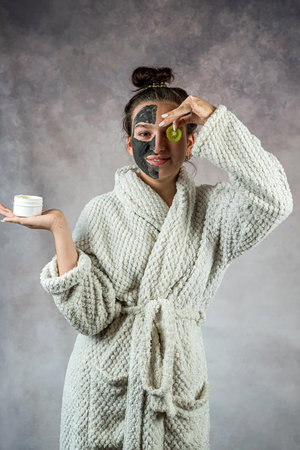 Young woman wear white bathrobe with holding kiwi fruit with black mask on her face. Skincare, self care or beautyの写真素材