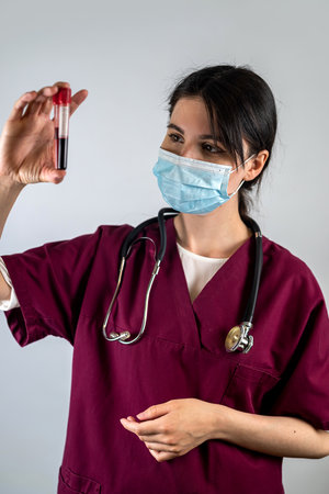 A nurse in a blue uniform wearing a mask holds a blood sample from a test tube for coronavirus in a laboratory for analysis isolated on a plain background. COVID pandemic outbreak concept. blood testの写真素材