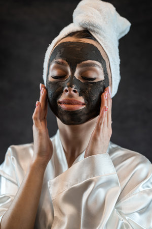 portrait of brunette caucasian woman wear white towel with black clay mud nourishing mask on face isolated on black. Skincare cleansing spa anti aging treatmentの写真素材