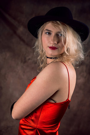 Beautiful blonde woman in red elegant evening dress and black hat posign in studio isolated on dark...