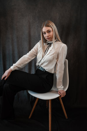 portrait of beautiful blonde woman in white blouse sitting on chair isolated on black, studioの写真素材