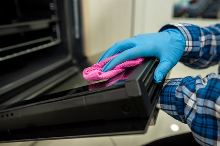 Female hands of a worker in protective gloves are cleaning and cleaning the oven with a rag and a...
