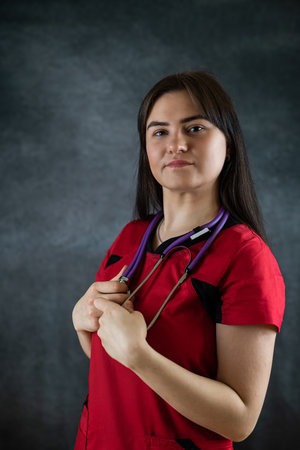 Woman hospital worker in red uniform with stethoscope looking at camera isolated on dark black background. Healthcare conceptの写真素材