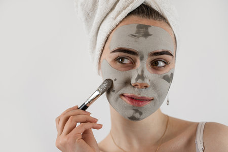 Pretty young lady putting clay black face mask with brush isolated on white. Cosmetic procedures conceptの写真素材