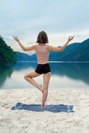 Young woman practices yoga at sand beach near lake with mountain background. Streching exercise in...
