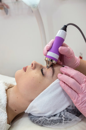 Young woman applying apparatus phonophoresis procedure on face. Hardware face cleaning procedure.