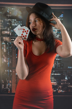 beautiful young girl in a red dress with a hat on her head holds house cards in her hands. a...