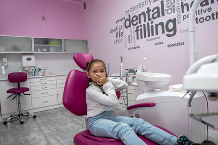 Close-up of a little girl in a dental chair sitting to do a dental procedure covering her mouth with her hand. child's fear of the dentistの写真素材