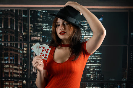 beautiful young girl in a red dress with a hat on her head holds house cards in her hands. a...
