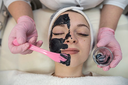 female lying at bad on spa clinic and receive cream black cosmetic mask. Clean skin, body care...