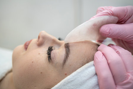 cosmetologist masseur doing faacial anti-agging procedure with gouache scraper for her female client. Treatment, body careの写真素材