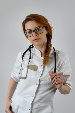 Portrait of young medical doctor in white uniform and stethoscope looking at camera on a grey background. Healthcare worker, general practitionerの写真素材