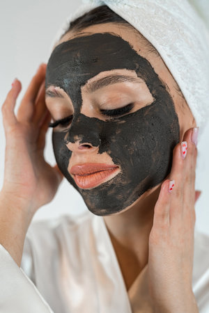 Young womanin white towel applying mud black mask on her face isolated on white, spa treatments at home. Perfect skin conceptの写真素材