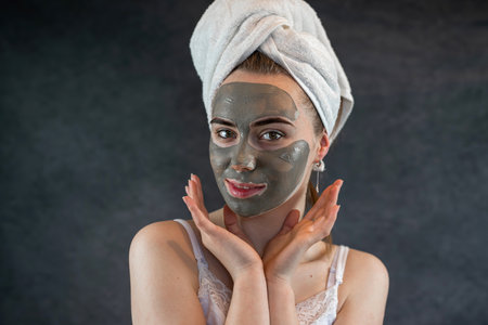 cute woman wear white towel with green nutritious clay facial mask look at camera isolated on black. Rejuvenation and skin care. Anti aging treatmentの写真素材
