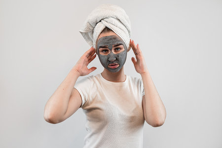 Pretty woman wear white t-shirt towel applying clay facial mask isolated on white. Face peeling mask, spa anti aging treatmentの写真素材
