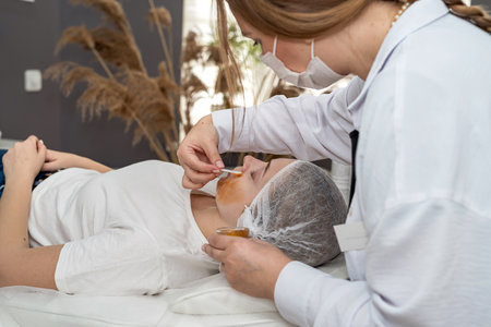 Cosmetologist applies yellow gold alginate mask with spatula on woman's face in spa salon, anti-aging cosmetic treatmentの写真素材