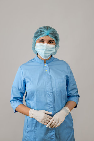 Portrait female doctor in blue uniform with protective medical gloves, mask cap isolated on grey. Medical staff, studio shotの写真素材