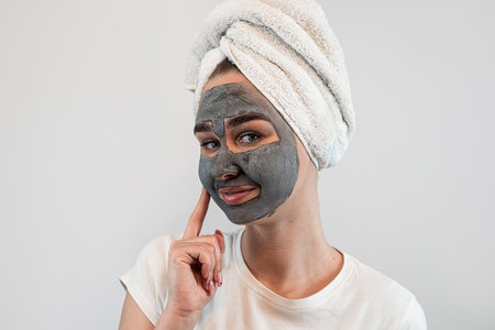 Attractive lady wear white towel applying black clay mask on her face isolated on white, skincare anti aging treatment, young woma enjoying wellnessの写真素材