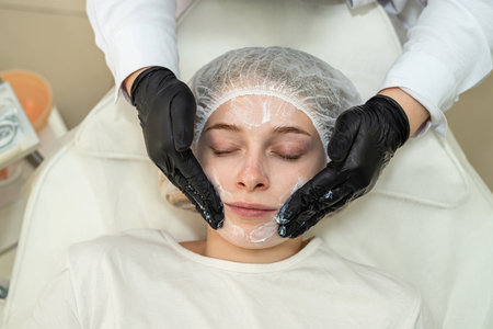 The cosmetologist applies nourishing white cream to the female patient's face at salon. Beauty anti wrinkle procedures conceptの写真素材
