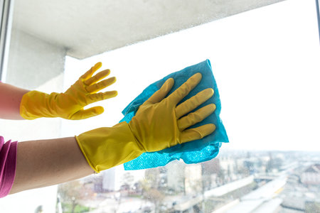 female hand wear yellow gloves with blue rag wipe window sill from dust at home. Cleaning concept
