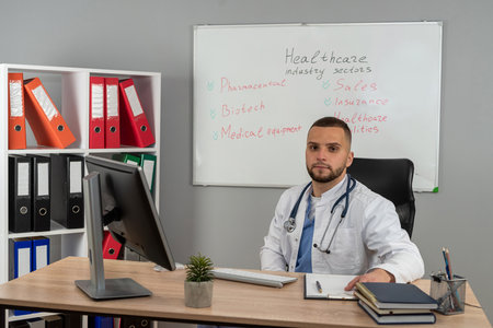 Caucasian young doctor wearing white uniform coat with stethoscope work at medical office, sitting at workdeskの写真素材