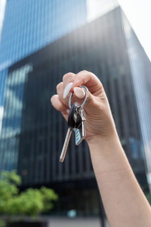 Woman hand holding new house key against the backdrop of urban development, new modern building....