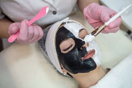 young woman with black and white clay cosmetic mask on half face lying a bad on spa salon. Skin care and treatment, lifestyleの写真素材