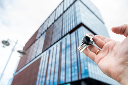 close up male hand holds house key against the backdrop of modern glass office center. Real estate agentの写真素材