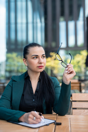 caucasian businessperson lady boss in a business dark suit holding a tablet looking away in big city on busy streetの写真素材