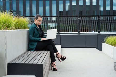 businesswoman or woman freelancer sitting outdoors with laptop sitting on bench cityscape in the...