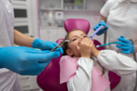 Pretty Caucasian child girl sitting on the couch looking scared with a drill in the hands of a professional female dentist in a modern children's clinic. a beautiful little child closes her mouth in fear. frightenedの写真素材