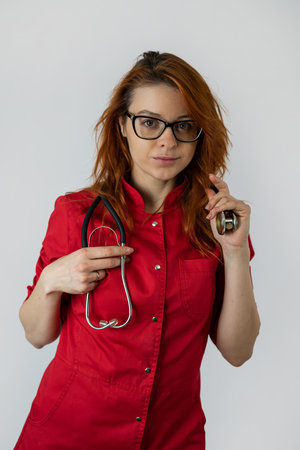 Caucasian female doctor in red uniform and stethoscope over light background. Portrait of health care workerの写真素材