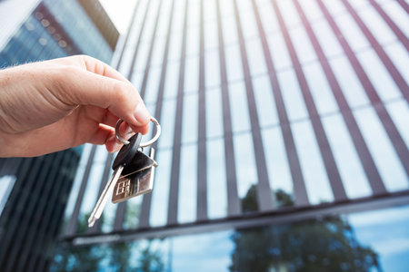 close up male hand holds house key against the backdrop of modern glass office center. Real estate agentの写真素材