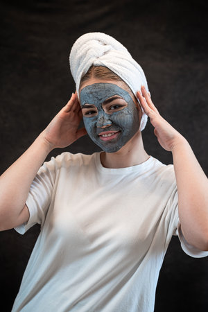 Young woman with a white towel applying cosmetic black mask of sea clay or mud isolated on black. Spa beauty treatment, skincare at home. Natural beauty and cosmetology conceptの写真素材