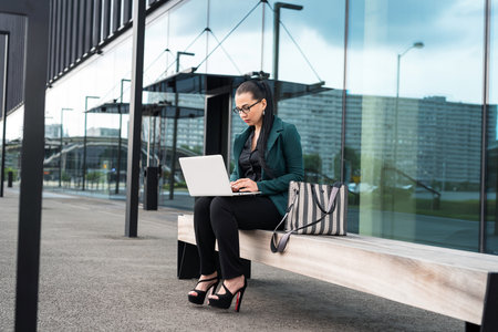 businesswoman or woman freelancer sitting outdoors with laptop sitting on bench cityscape in the backgroundの写真素材