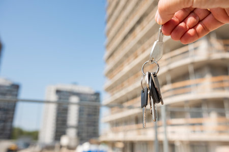 A person is standing upright while holding a set of keys, positioned in front of a tall and imposing building that stretches high towards the sky above themの写真素材