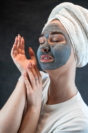 Young woman with a white towel applying cosmetic black mask of sea clay or mud isolated on black. Spa beauty treatment, skincare at home. Natural beauty and cosmetology conceptの写真素材