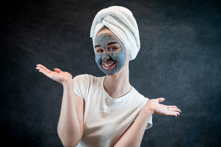 Young woman with a white towel applying cosmetic black mask of sea clay or mud isolated on black. Spa beauty treatment, skincare at home. Natural beauty and cosmetology conceptの写真素材