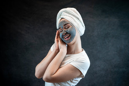 Young woman with a white towel applying cosmetic black mask of sea clay or mud isolated on black. Spa beauty treatment, skincare at home. Natural beauty and cosmetology conceptの写真素材