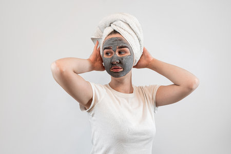 young caucasian woman in towel on head with black clay or mud facial mask isolated on white. Anti aging treatment at home. Face peeling maskの写真素材