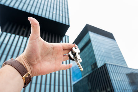 close up male hand holds house key against the backdrop of modern glass office center. Real estate agentの写真素材