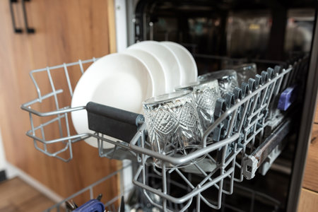 Open integrated dishwasher machine with clean cutlery glasses, dishes. Built-in dishwasher in modern...