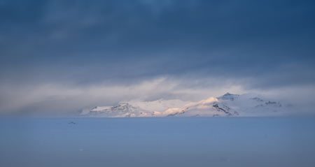 Focssed morning sunshine on a mounting range, winter day in Iceland with overcast sky and snow-covered landの写真素材