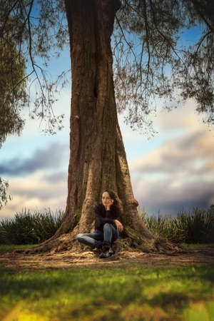 Vertical view where a young woman is shown in the foreground sitting in the shade of a very large tree and in the background a very beautiful skyの写真素材