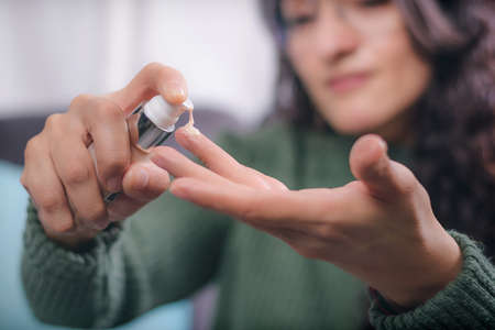 Concept of care and beauty. Hands of young woman holding a bottle of cosmetic, the woman is sitting in an armchair at home and pours the cosmetic content in her hand to apply on her face.の写真素材