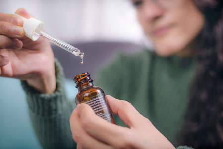 Concept of care and beauty. Hands of young woman holding a container with dropper showing the content, the woman is sitting in an armchair at home.の写真素材