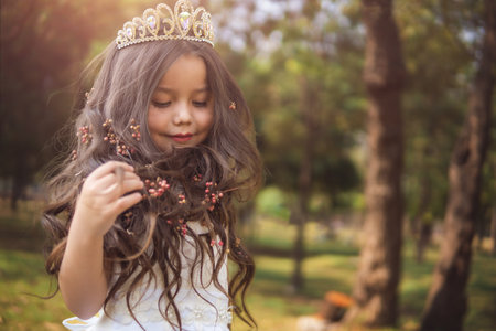 Little girl in white dress and princess crown, she plays with her long blonde hair, copy space, children's day theme.の写真素材