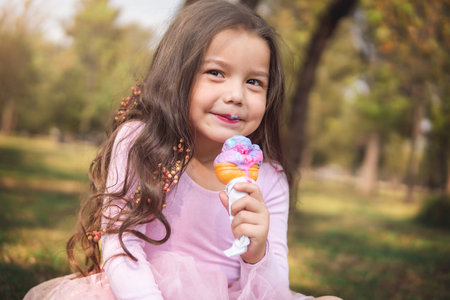 Cute blonde curly haired girl eating a nice ice cream in the forest on a nice summer day, she looks at the camera, children's day concept, happy girl enjoying a hot day.の写真素材