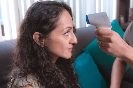 A man measures a woman's temperature with a digital infrared thermometer at home sitting on the couch, The image highlights health and safety protocols in action.の写真素材